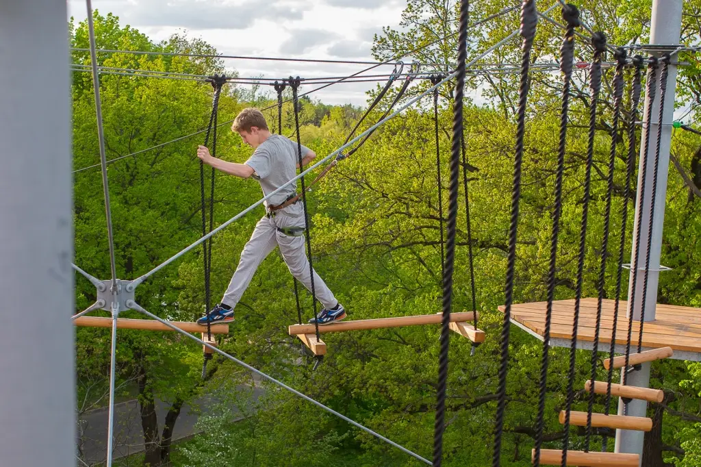 A young boy in a safety harness reaching for the final hold at the top of a tall outdoor rope climbing tower.