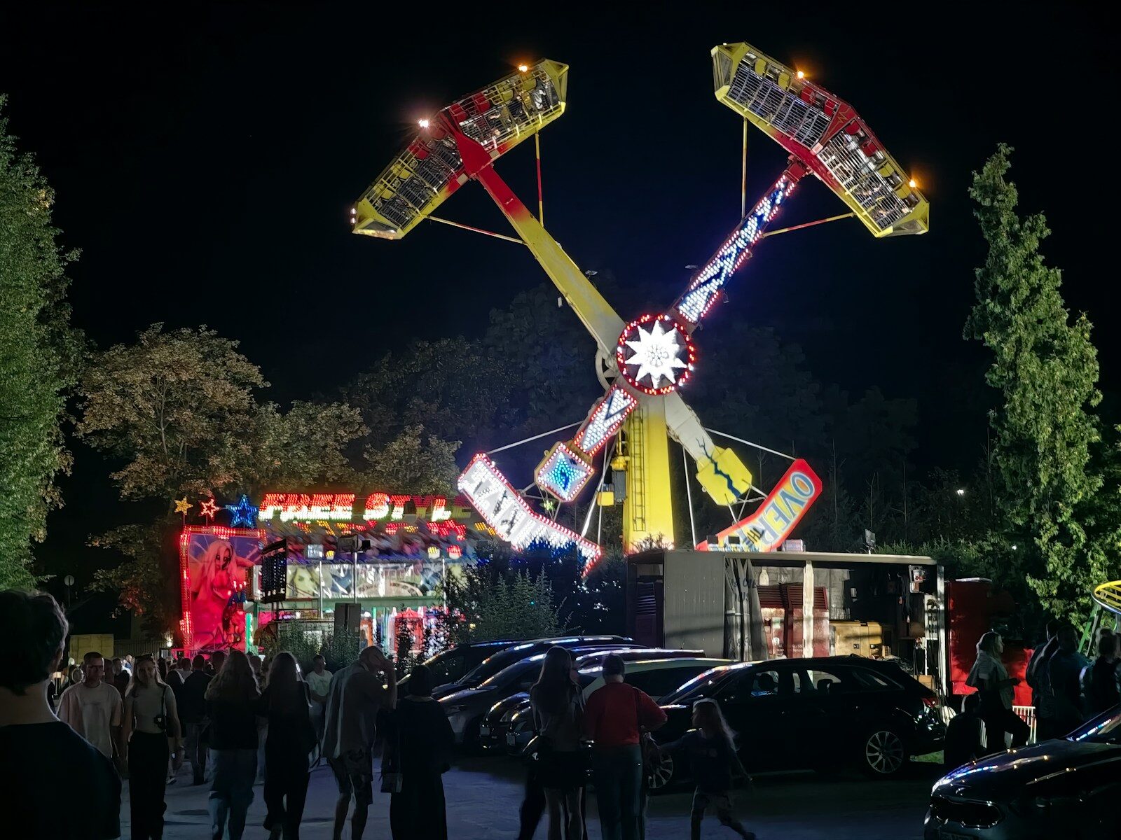 A crowd of people walking past a brightly lit carnival ride at night