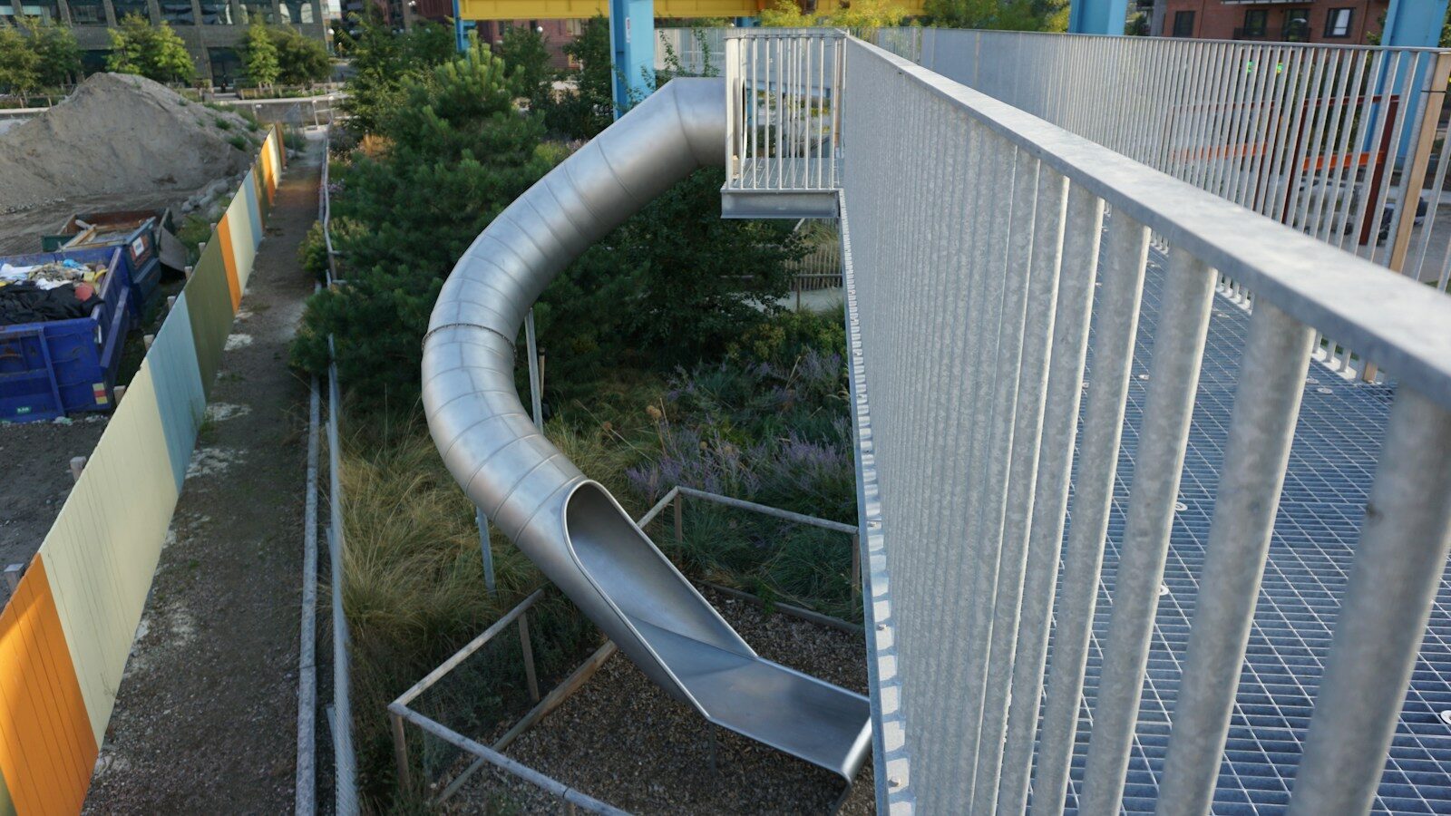 A metal tube slide descending from an elevated walkway into a garden