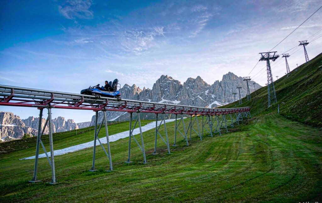 mountain coaster running down a green summer ski slope with snowy peaks in the background